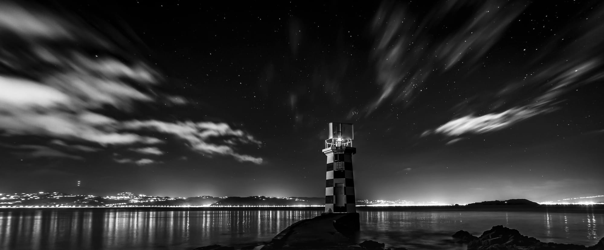 A lighthouse in a starry night, watching over an inlet to a distant town, lightening the horizon.