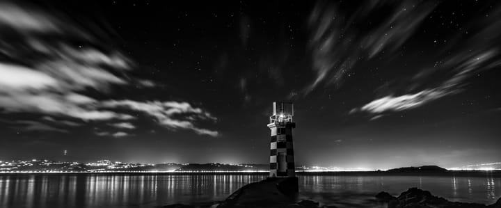 A lighthouse in a starry night, watching over an inlet to a distant town, lightening the horizon.